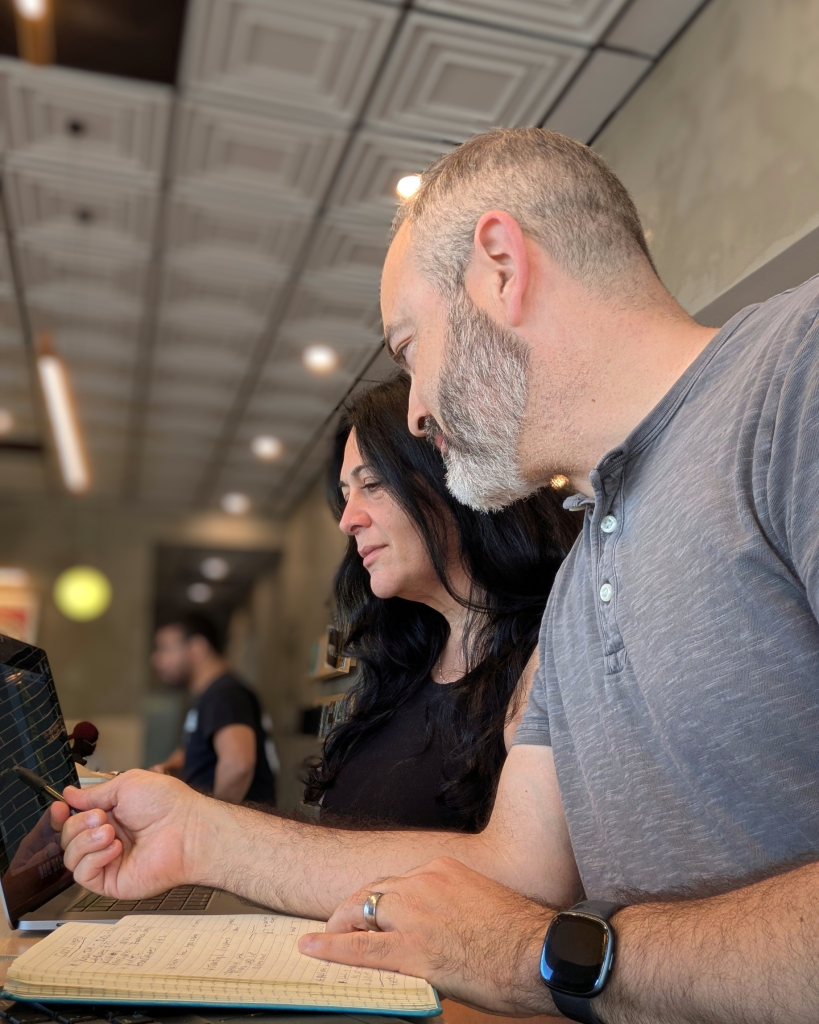 A man with a gray beard points at a laptop next to a woman with black hair.
